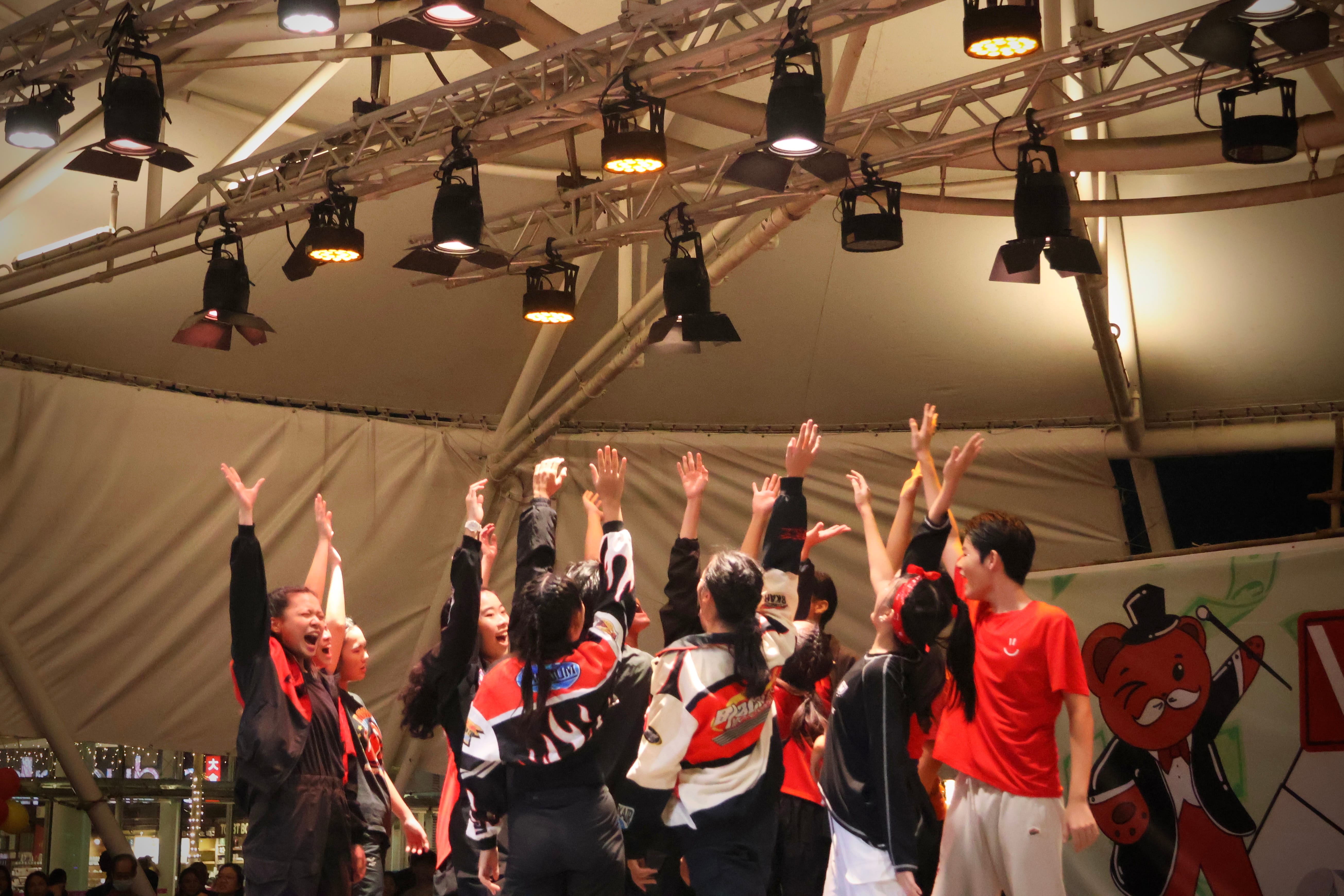 A group of performers in red and black outfits raise their hands on a lit indoor stage with overhead spotlights.