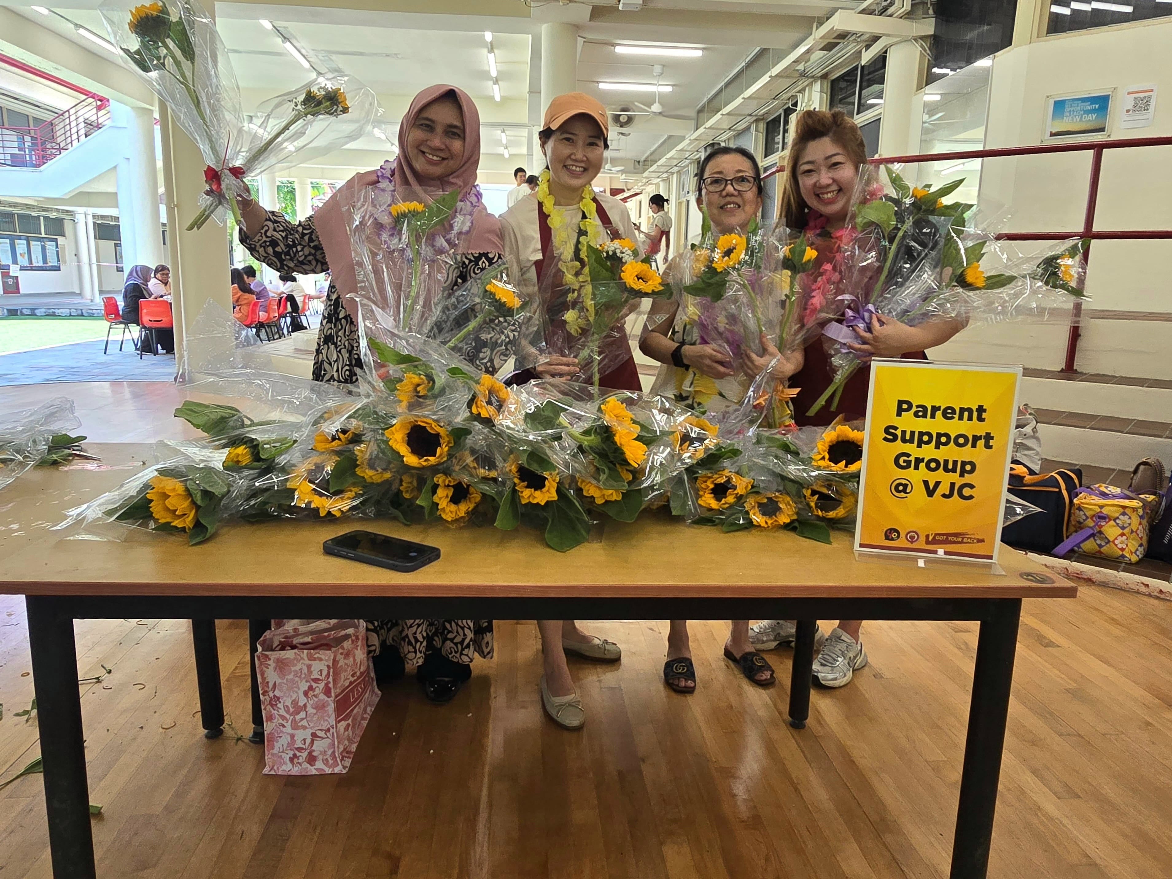 Four smiling women hold sunflower bouquets behind a table at a Parent Support Group at VJC event.