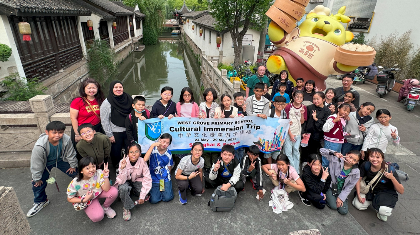 A large group of students and teachers pose by a canal in China, holding a West Grove Primary School Cultural Immersion Trip banner.