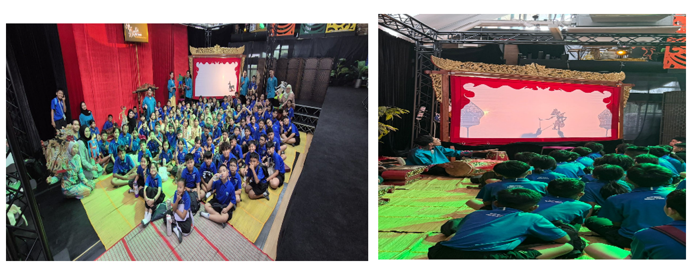 Children in blue uniforms sit on mats watching a shadow puppet show on a lit screen framed by a decorative stage.