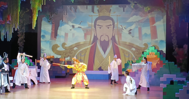 Performers in traditional Chinese costumes on stage with a large animated emperor backdrop and colorful scenery.