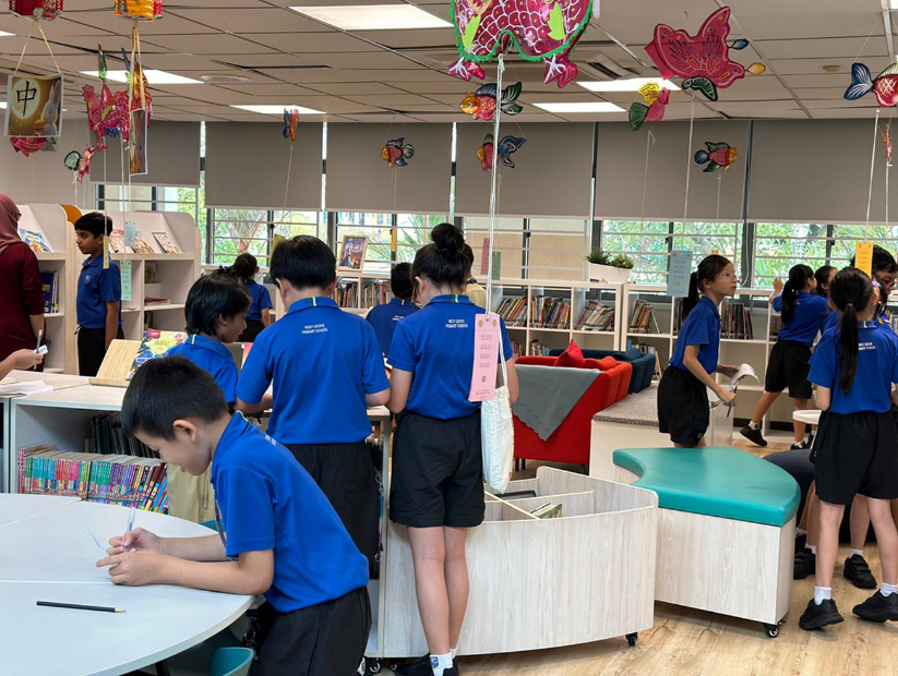 Students in blue uniforms browse a decorated school library with colorful hanging ornaments and bookshelves.
