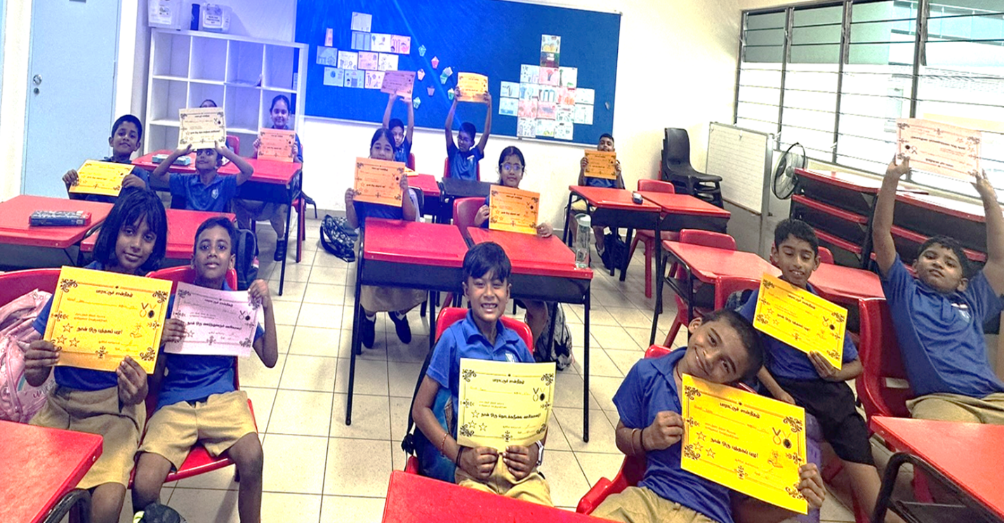 Children in blue uniforms sit at red desks in a classroom, smiling and holding colorful award certificates.