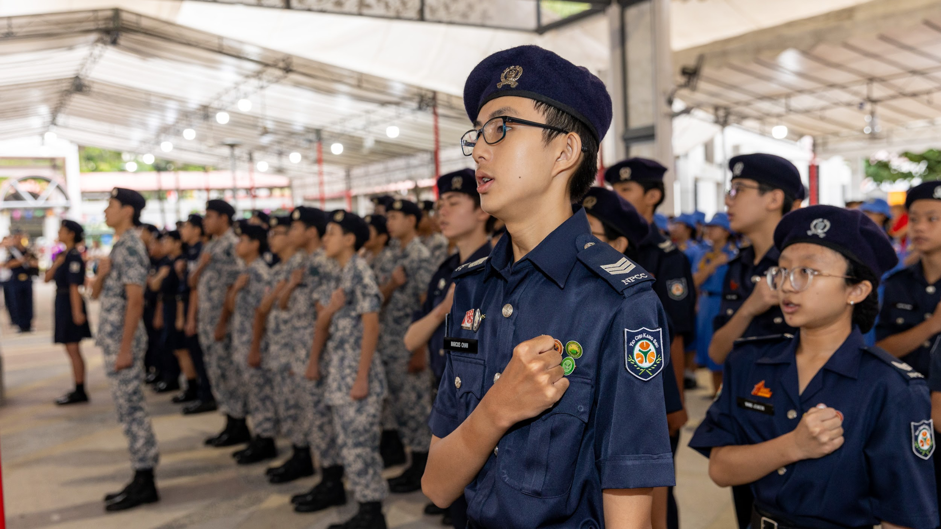 Young cadets in NPCC uniforms and berets stand at attention with fists raised during a formal parade under a tent.