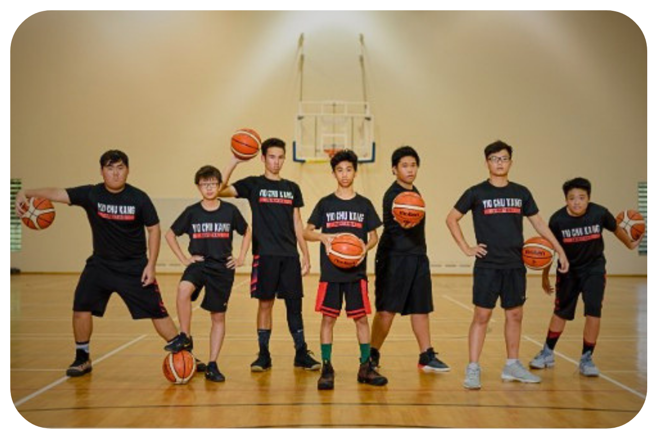 Seven young players in black jerseys pose with basketballs on an indoor court.