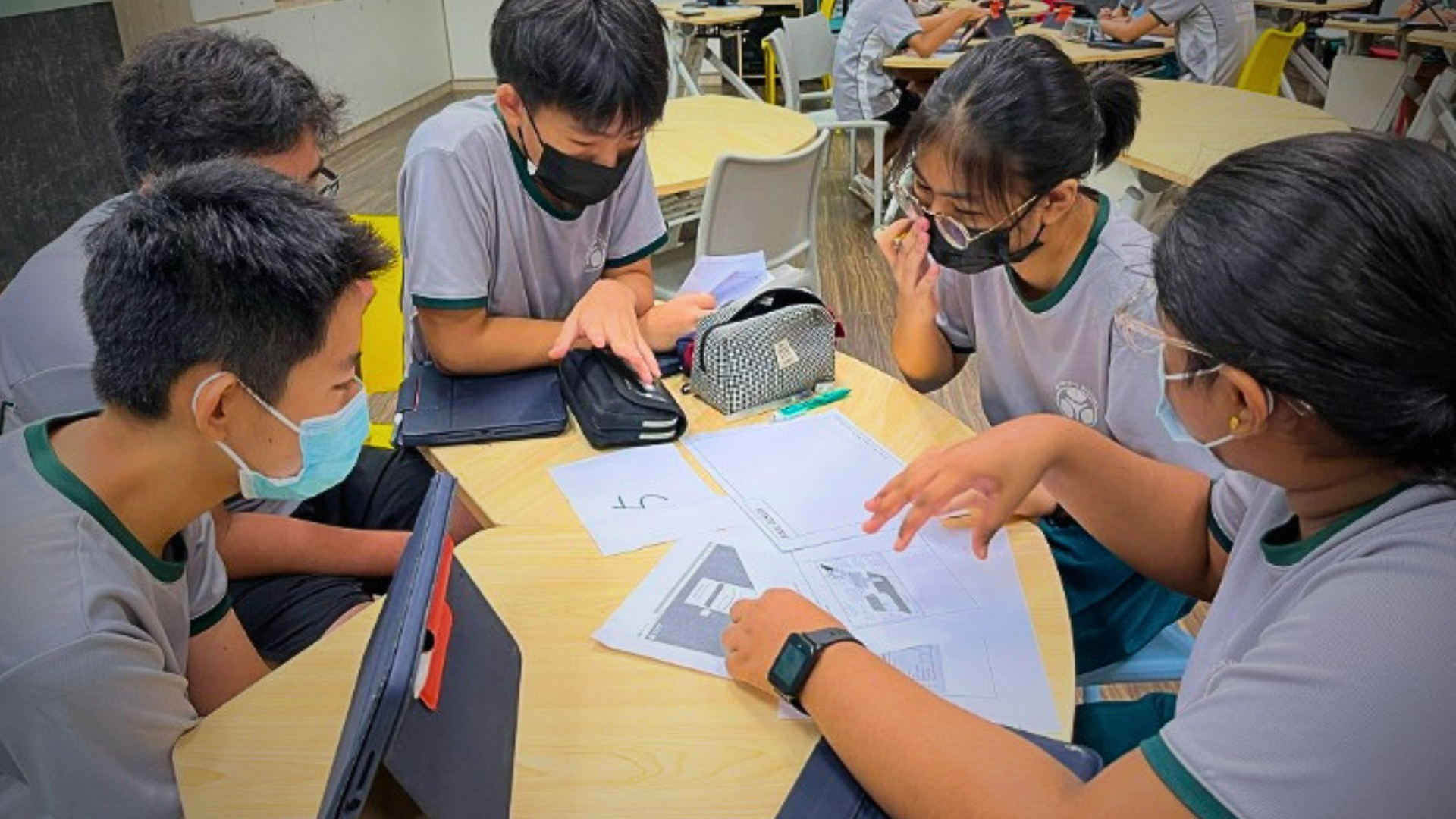 Seven students in uniforms pose around a wooden picnic table with project displays, posters visible on the wall behind them.