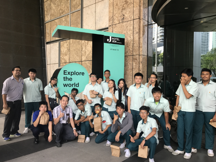 Group of students in light green uniforms posing outside Hotel Jen beside an "Explore the world" sign.