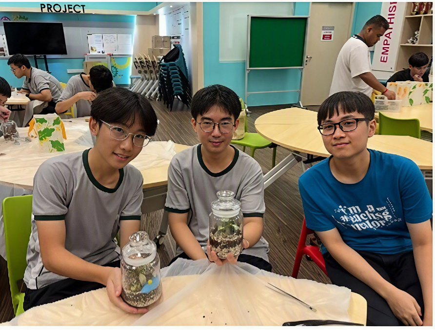 Three students seated at a workshop table displaying their handmade glass jar terrariums, with a classroom workspace in the background.