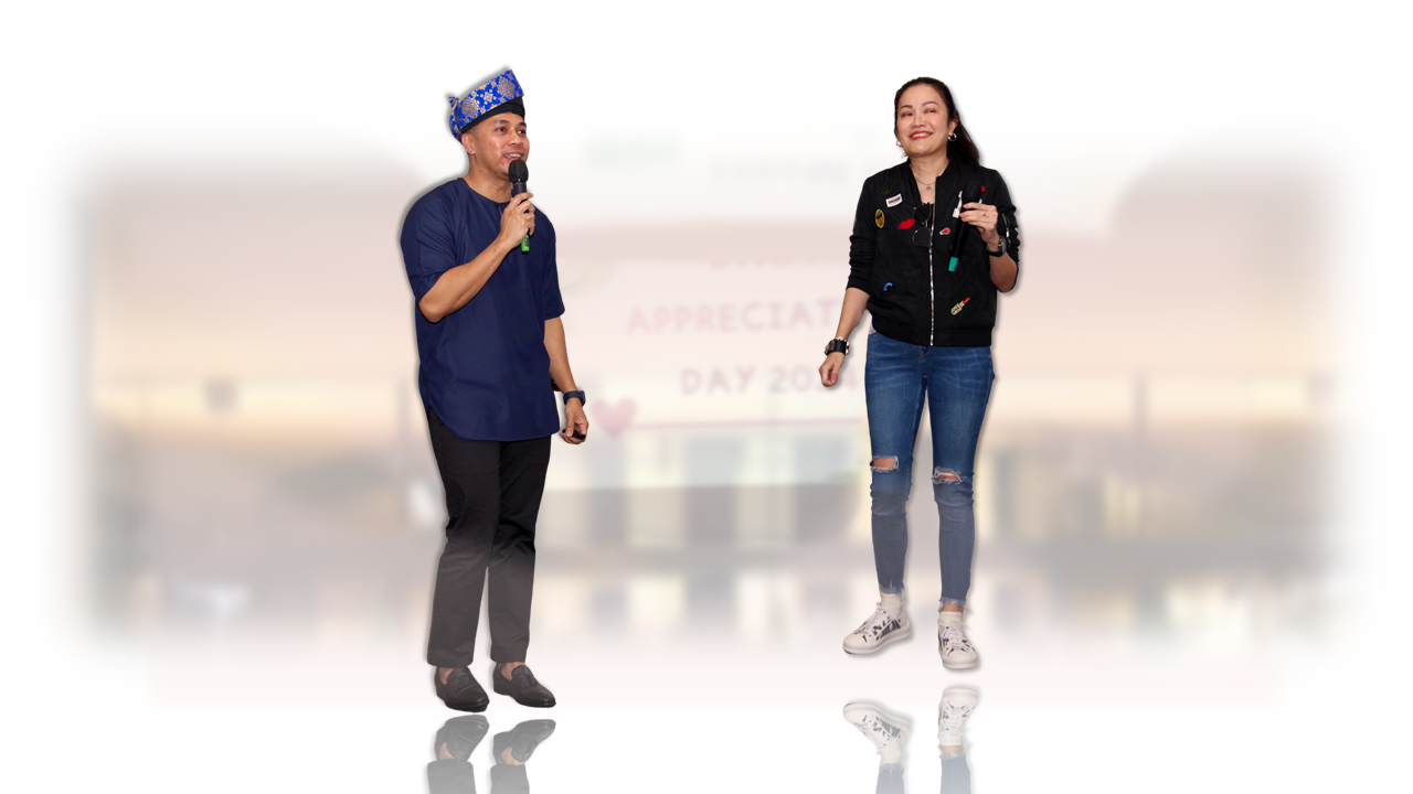 Man in blue shirt and decorative cap holds microphone beside smiling woman in black jacket and jeans at an Appreciation Day event.