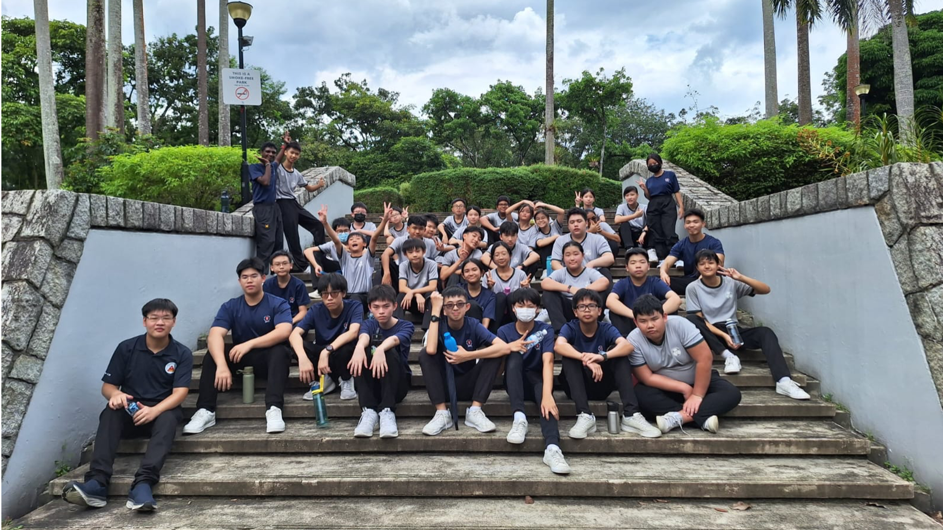 A large group of students in navy and grey uniforms posing on outdoor stone steps surrounded by green trees.