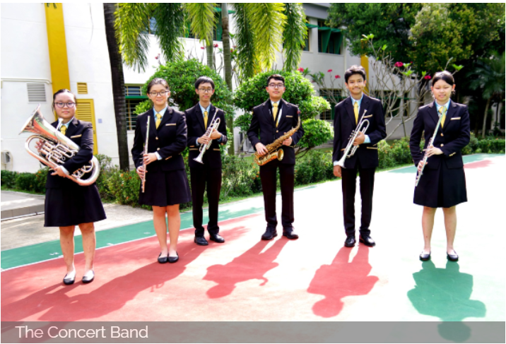 Six students in black uniforms hold brass instruments outdoors on a colorful court surrounded by trees.
