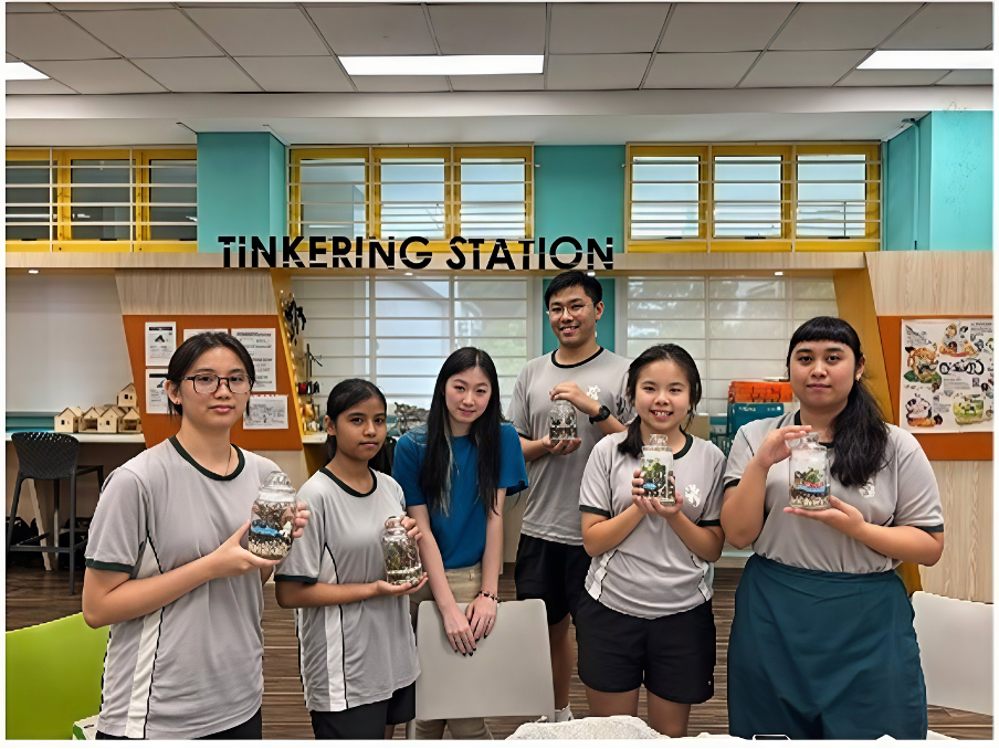 Enter a descriptive alt text.A group of students standing together in front of a "TINKERING STATION" sign in a school maker space, holding up glass jar terrariums they crafted.