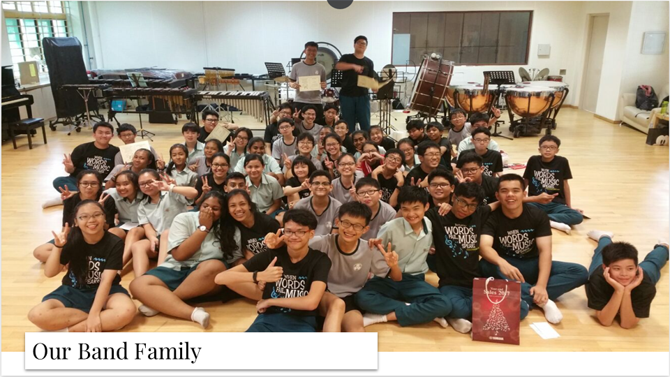 Large group photo of band members sitting on the floor of a music practice room surrounded by percussion instruments.