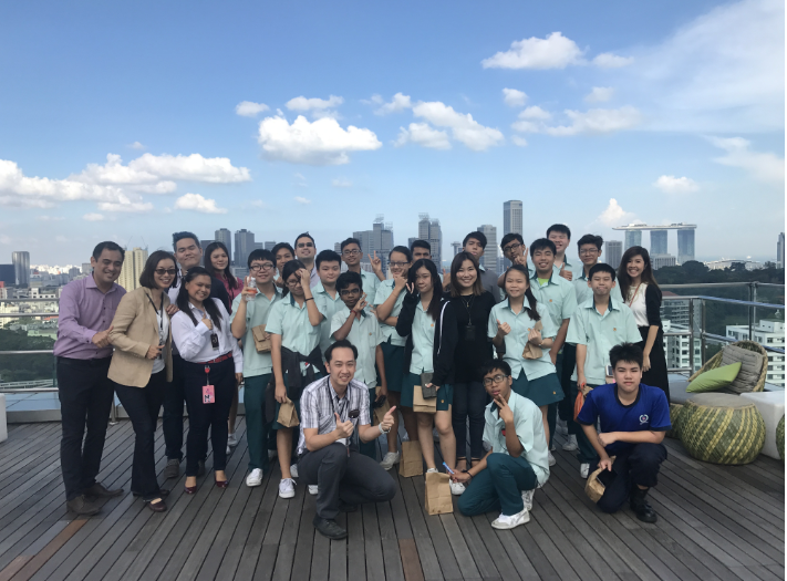 A large group of students in teal uniforms pose on a rooftop deck with a city skyline behind them.