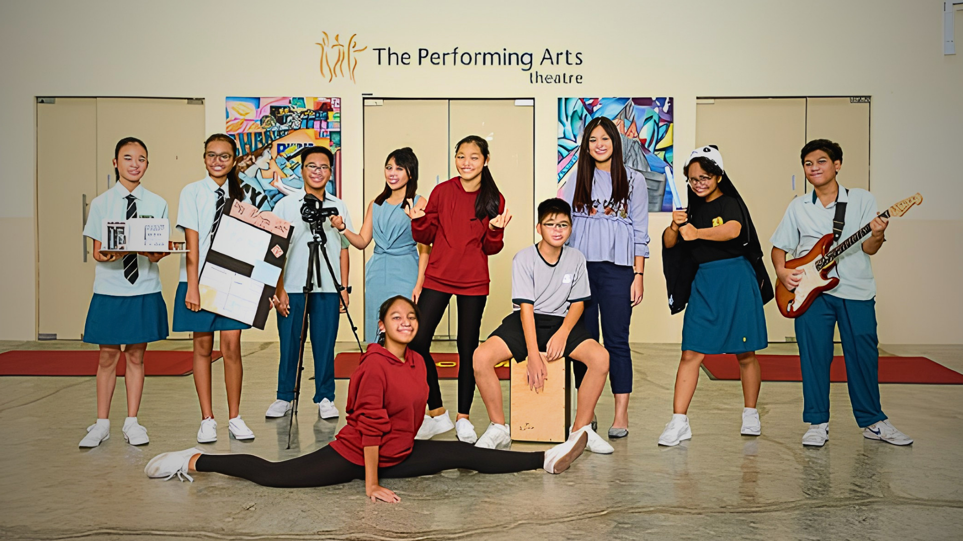 Ten students pose outside The Performing Arts Theatre, one doing splits, another holding a guitar, with colorful artwork behind them.