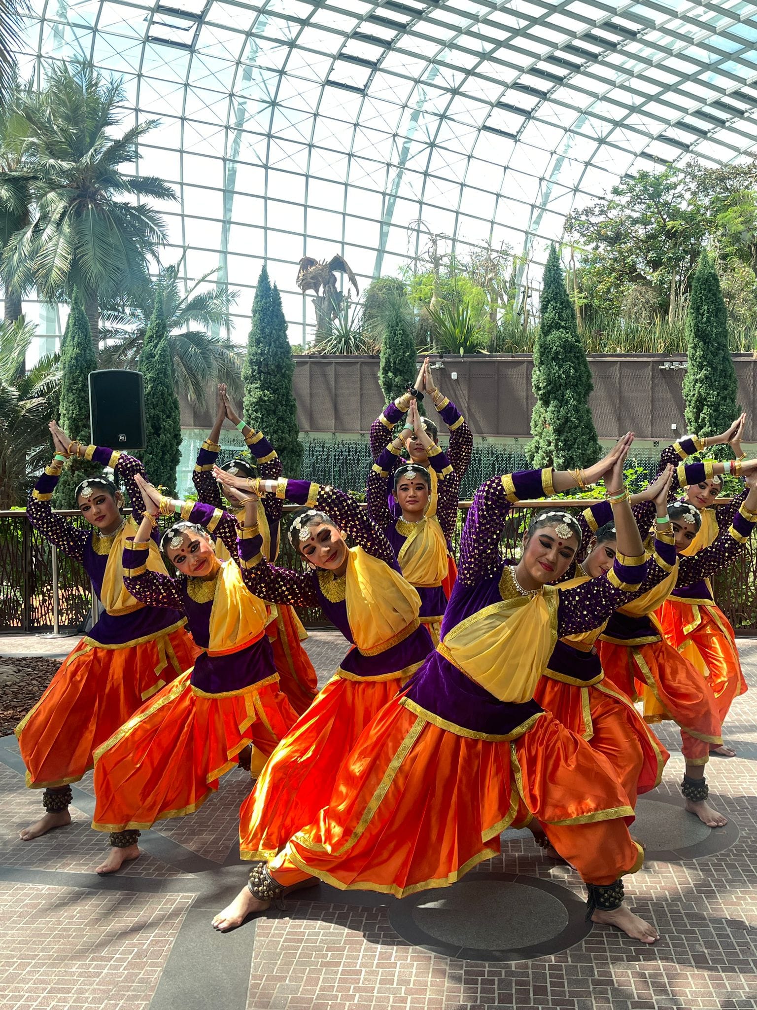 Group of female Indian classical dancers in orange and purple costumes performing inside a glass-domed greenhouse.