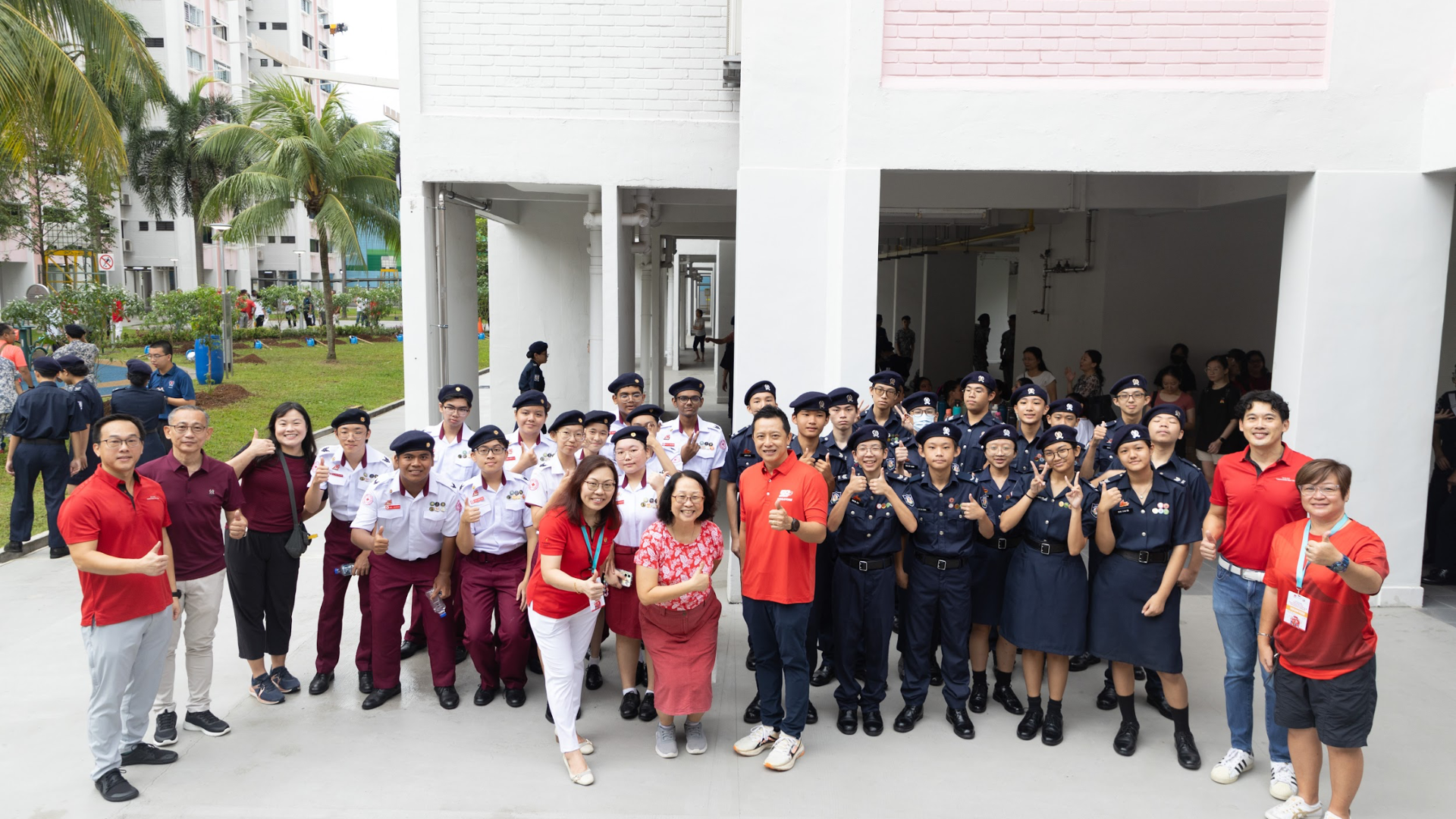 A large group of uniformed youth cadets and adults in red shirts pose together outdoors near a white building.