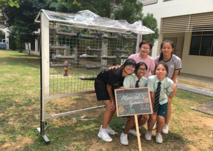 Five people pose outdoors by a metal frame structure, holding a chalkboard sign reading "Mendel's Garden 2.0."