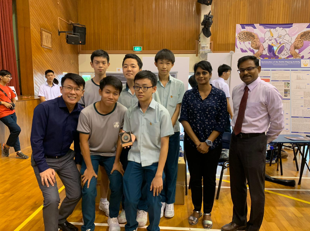 A group of students and two adult teachers pose together in a school hall near research project display boards.