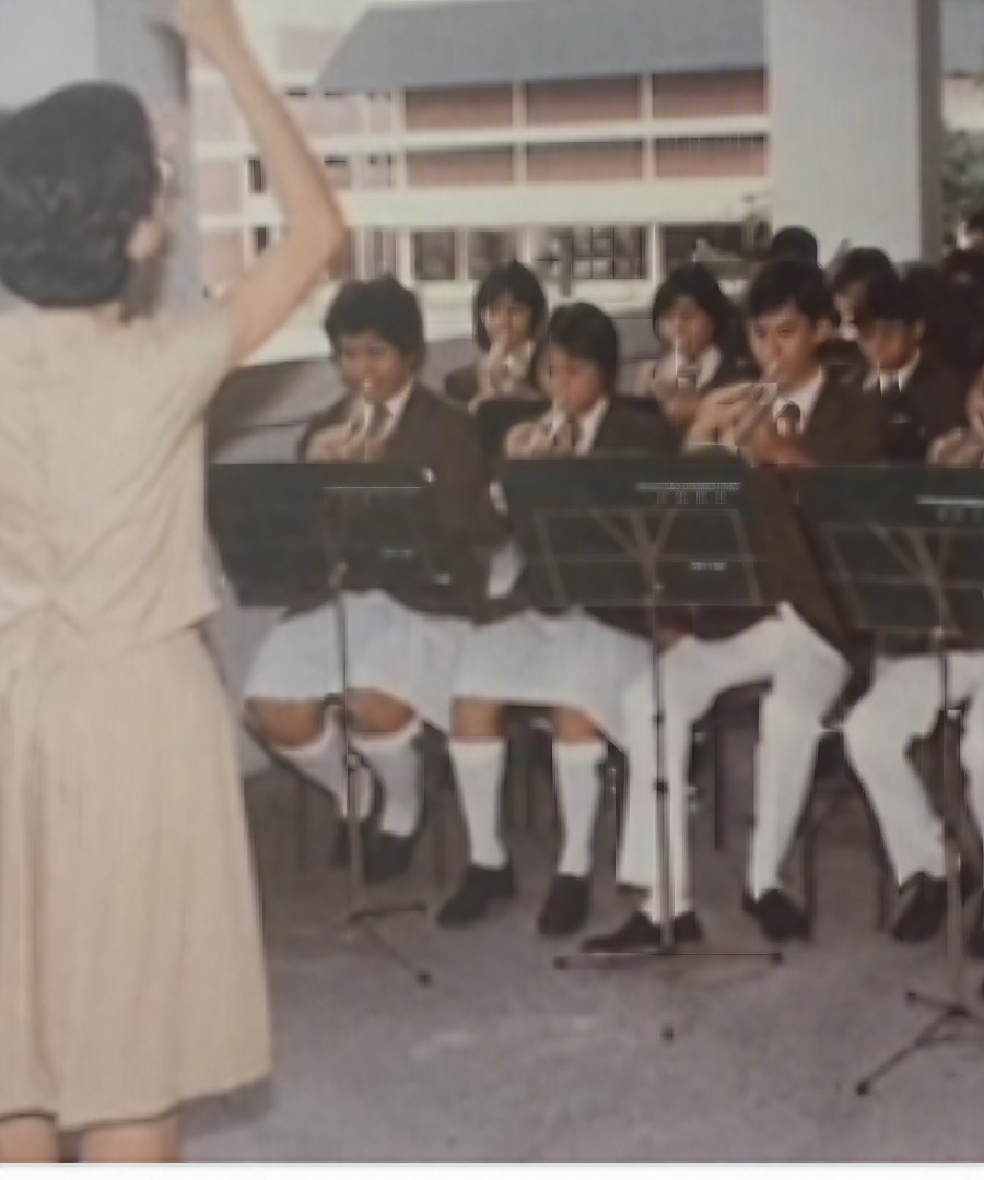 A instructor conducting a seated student musical ensemble performing with wind instruments under a covered outdoor concourse.