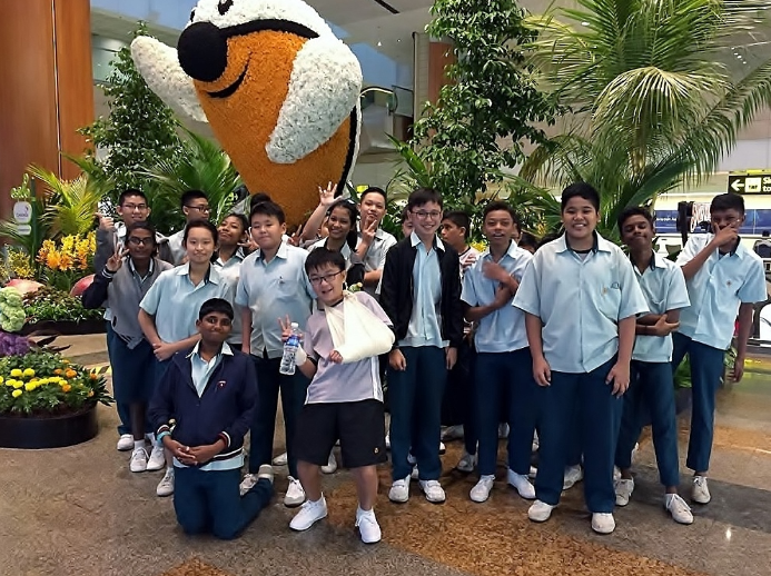 Group of students in light blue uniforms posing in front of a large floral bird sculpture near tropical plants.