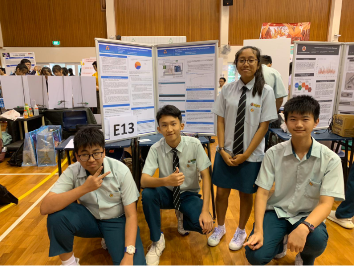 Four students in school uniforms pose in front of research poster boards labeled E13 at a science fair.