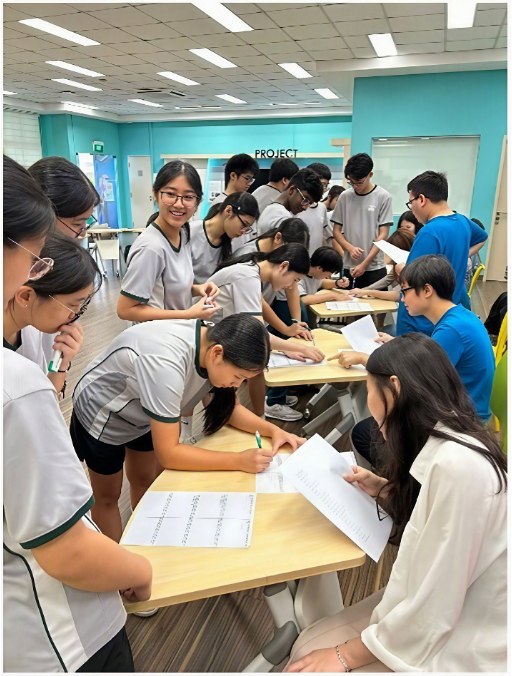 Students collaborating in an active classroom environment, leaning over grouped desks to complete worksheets alongside facilitators.