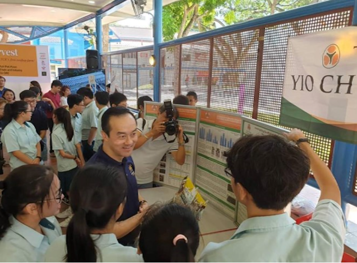 A smiling man poses with students in school uniforms at an outdoor exhibition near a Yio Chu banner.