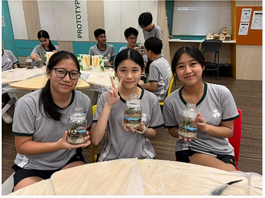 Three female students sitting at a round table smiling while holding up their completed glass jar terrariums in a school activity room.