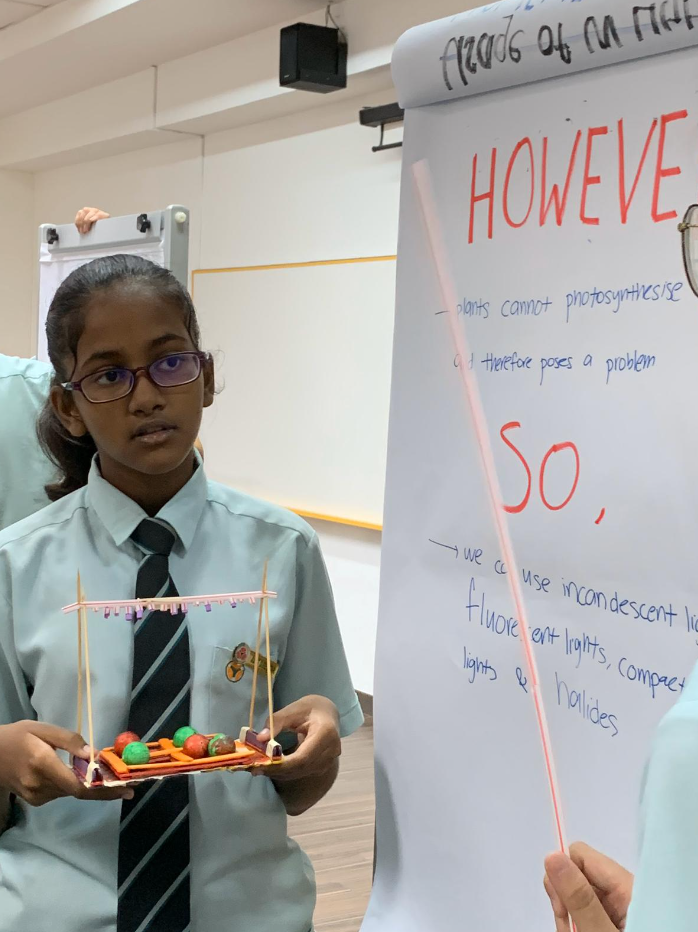 A uniformed schoolgirl with glasses holds a small model while standing next to a flipchart about photosynthesis.