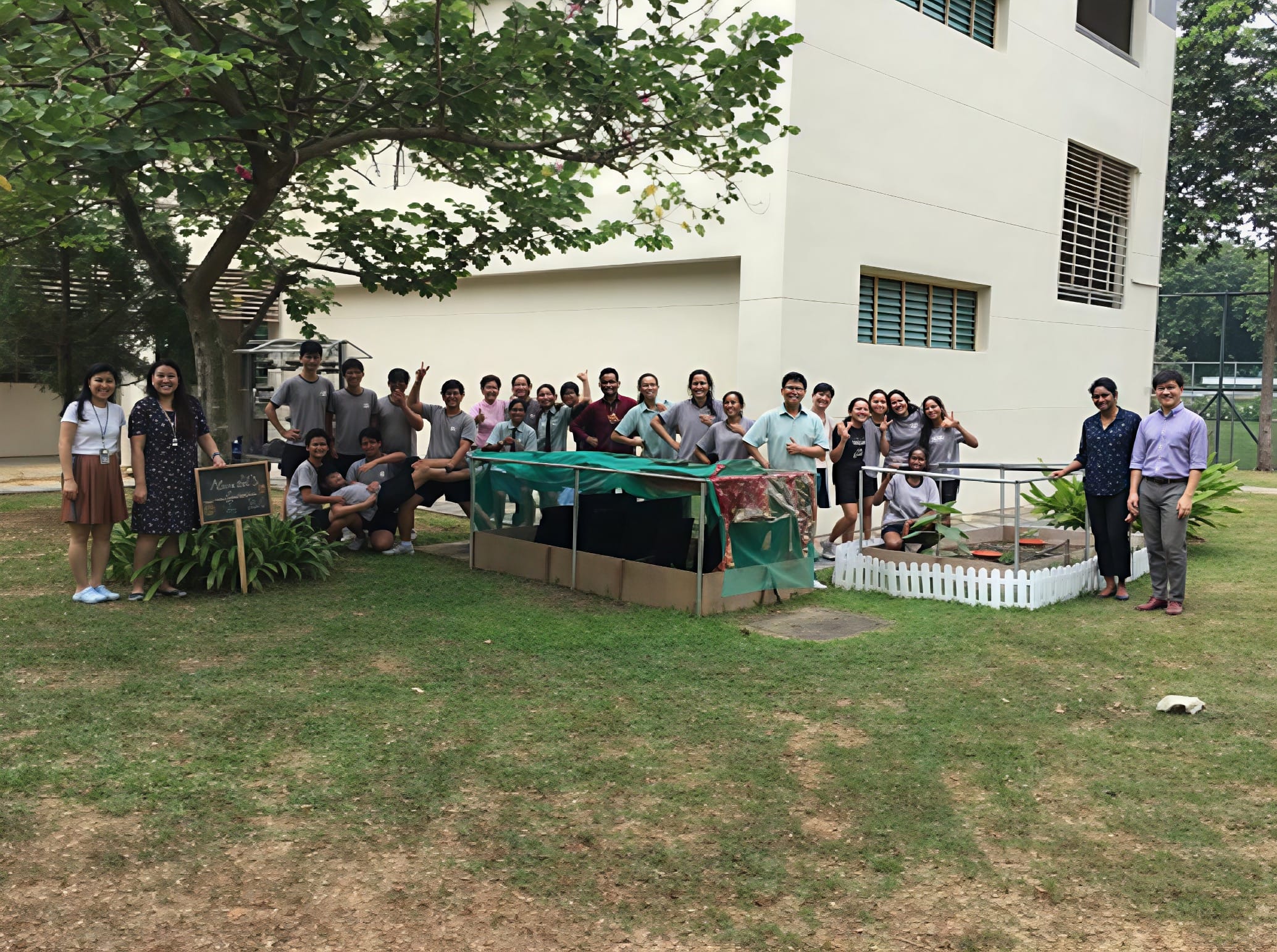 A large group of students and teachers pose outdoors on grass near a white building with a netted garden bed.