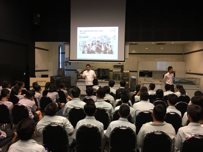 A presenter in a white chef's coat addresses a large seated audience in a culinary classroom with a projected slide.