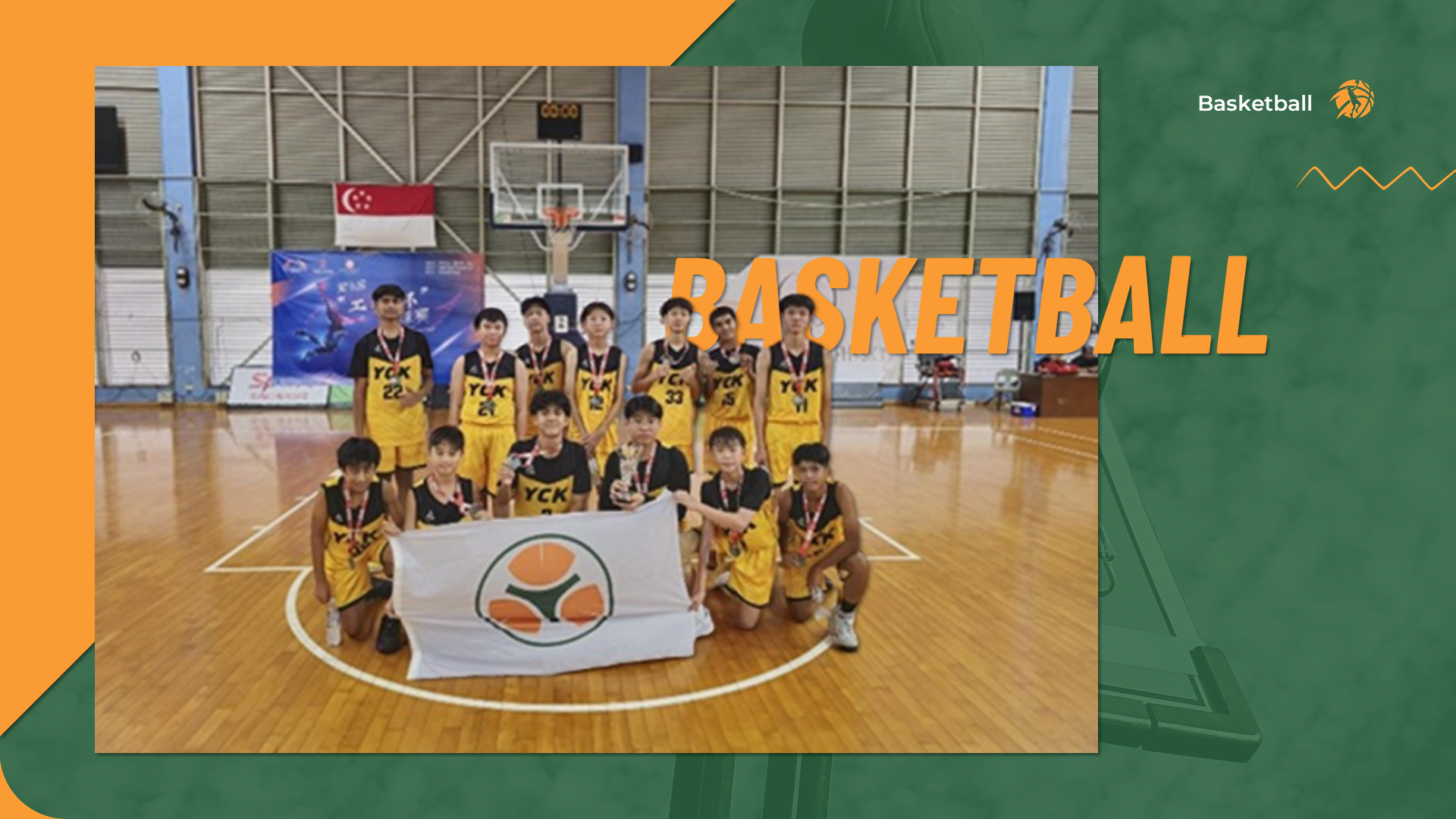Victory team photo of basketball players holding a trophy, medals, and school flag on the court.