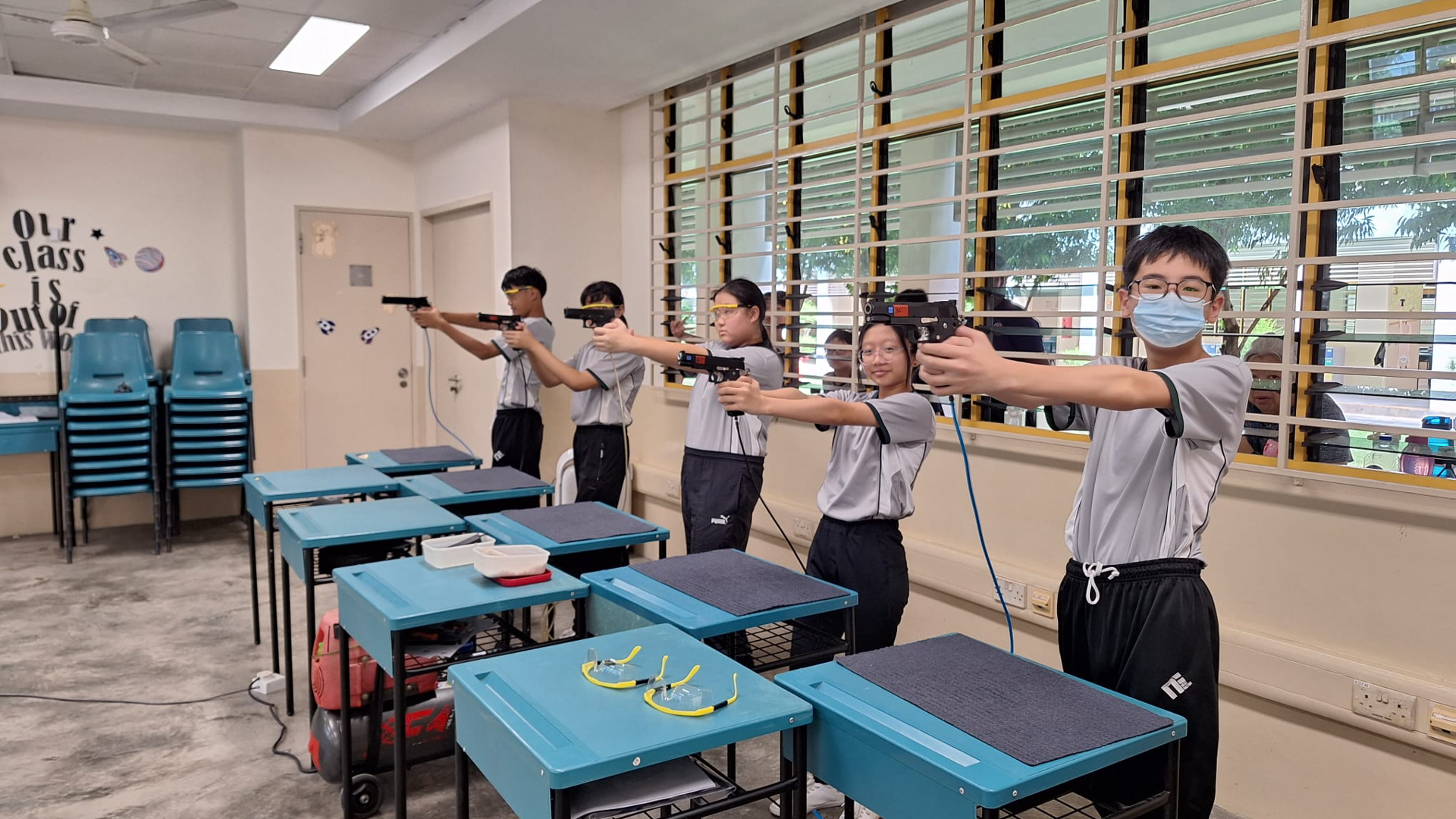 Four students in a classroom aim air pistols in a row, wearing safety goggles, beside blue school desks.