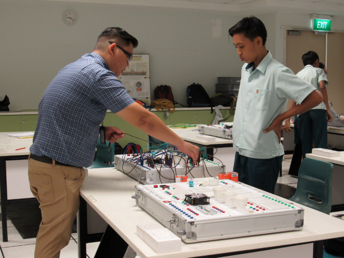 A teacher and student work with an electronics training board with wires and buttons in a classroom.