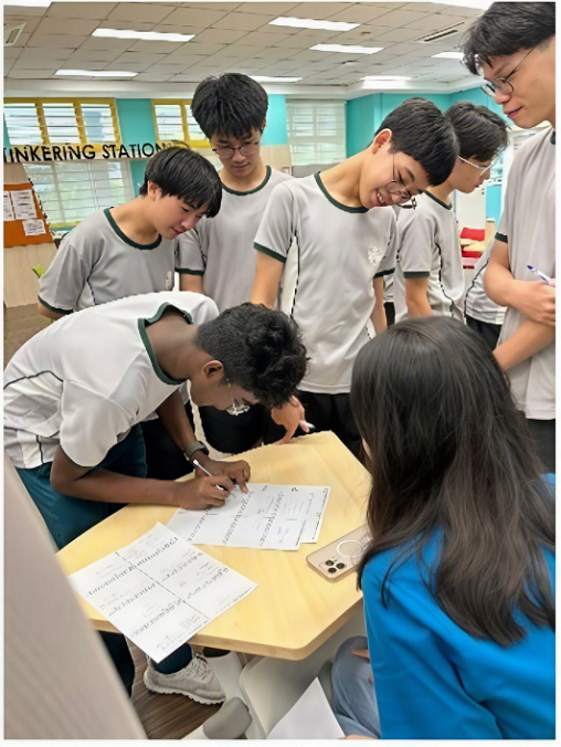 A group of male students leaning over a modular desk in a classroom to watch a classmate fill out a worksheet during a group problem-solving activity.