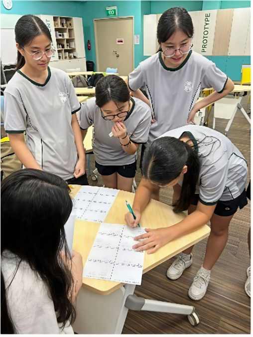 Four female students standing around a small desk, watching attentively as one classmate writes answers on a worksheet.