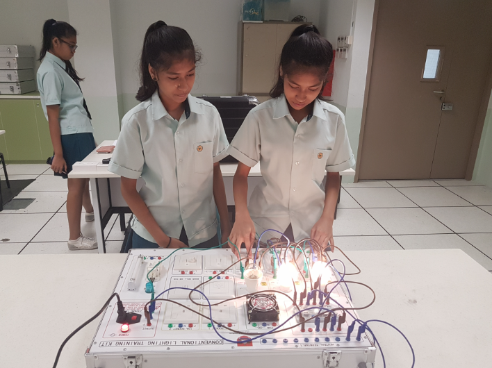 Two school girls in uniforms work on a conventional lighting training kit with wires and lit bulbs on a table.