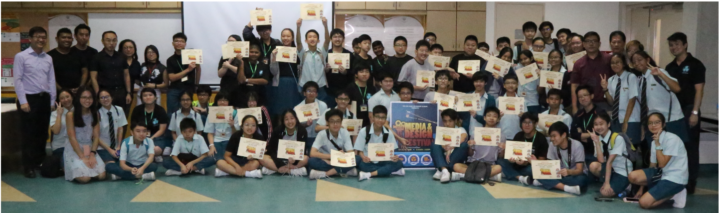 A wide group photograph of students and teachers posing together on the floor while holding up certificates of achievement around the 2018 Media & Design Festival banner.