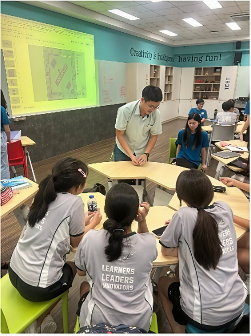 Students seated around modular desks listening to an instructor demonstrate a group activity, with a board game presentation projected on the wall behind them.