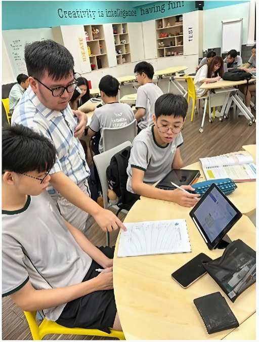 A teacher leaning over to guide two seated students through an exercise on a notebook and tablet.