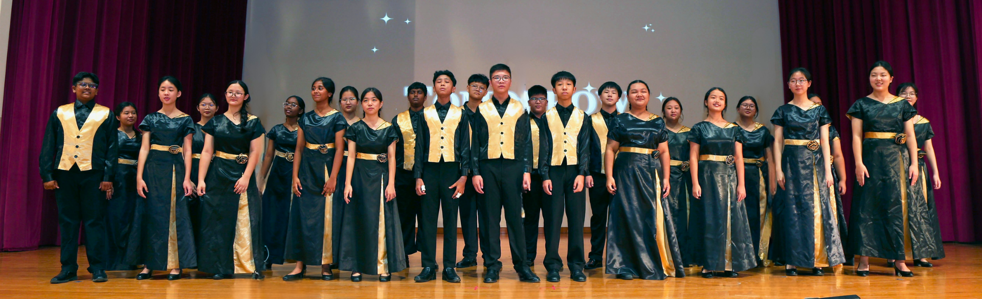 Panoramic photo of student choir members dressed in black and gold formal attire performing on a school stage.