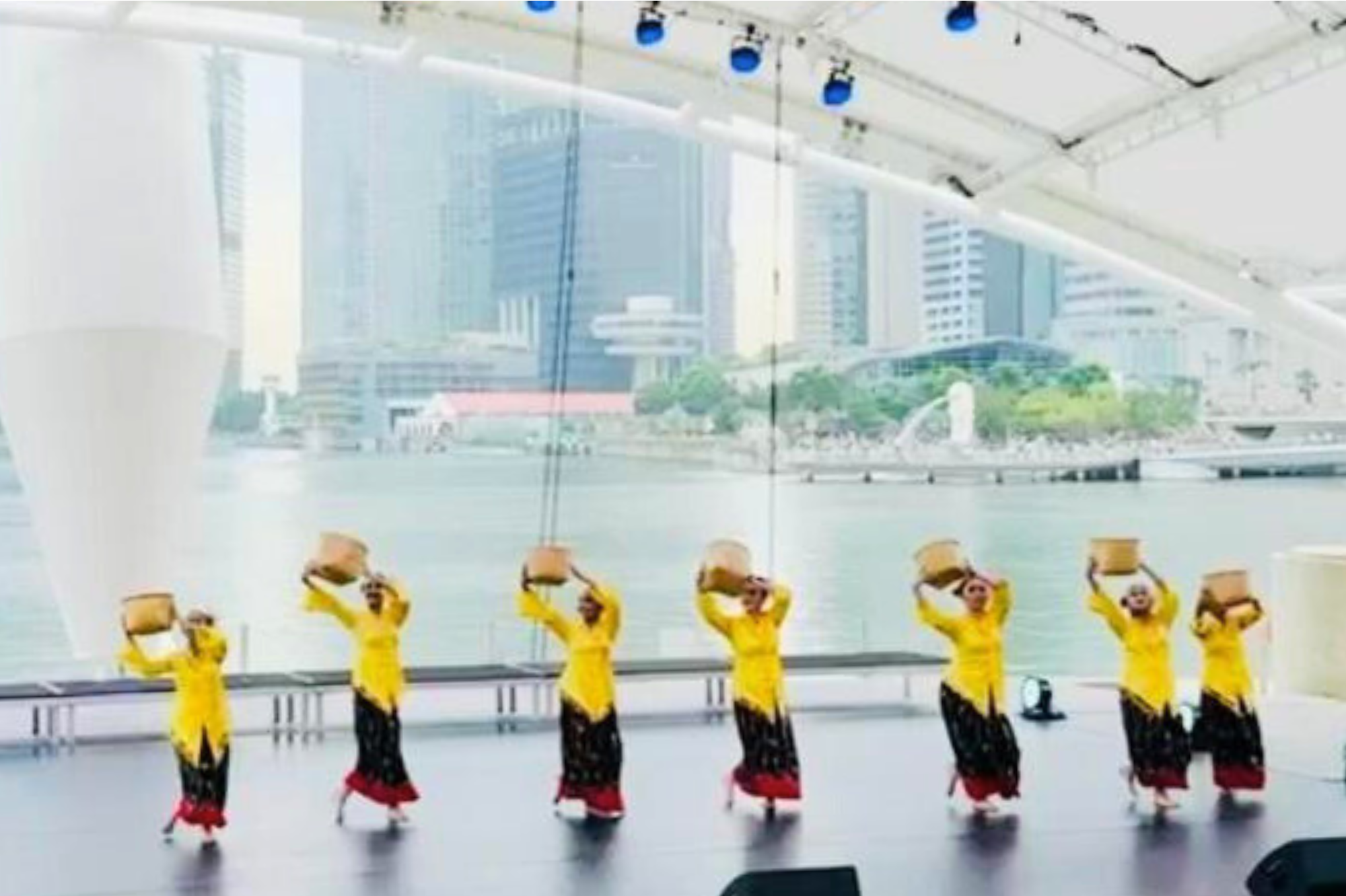 Dancers in yellow and black traditional costumes balance baskets on their heads on an indoor stage overlooking a city waterfront.