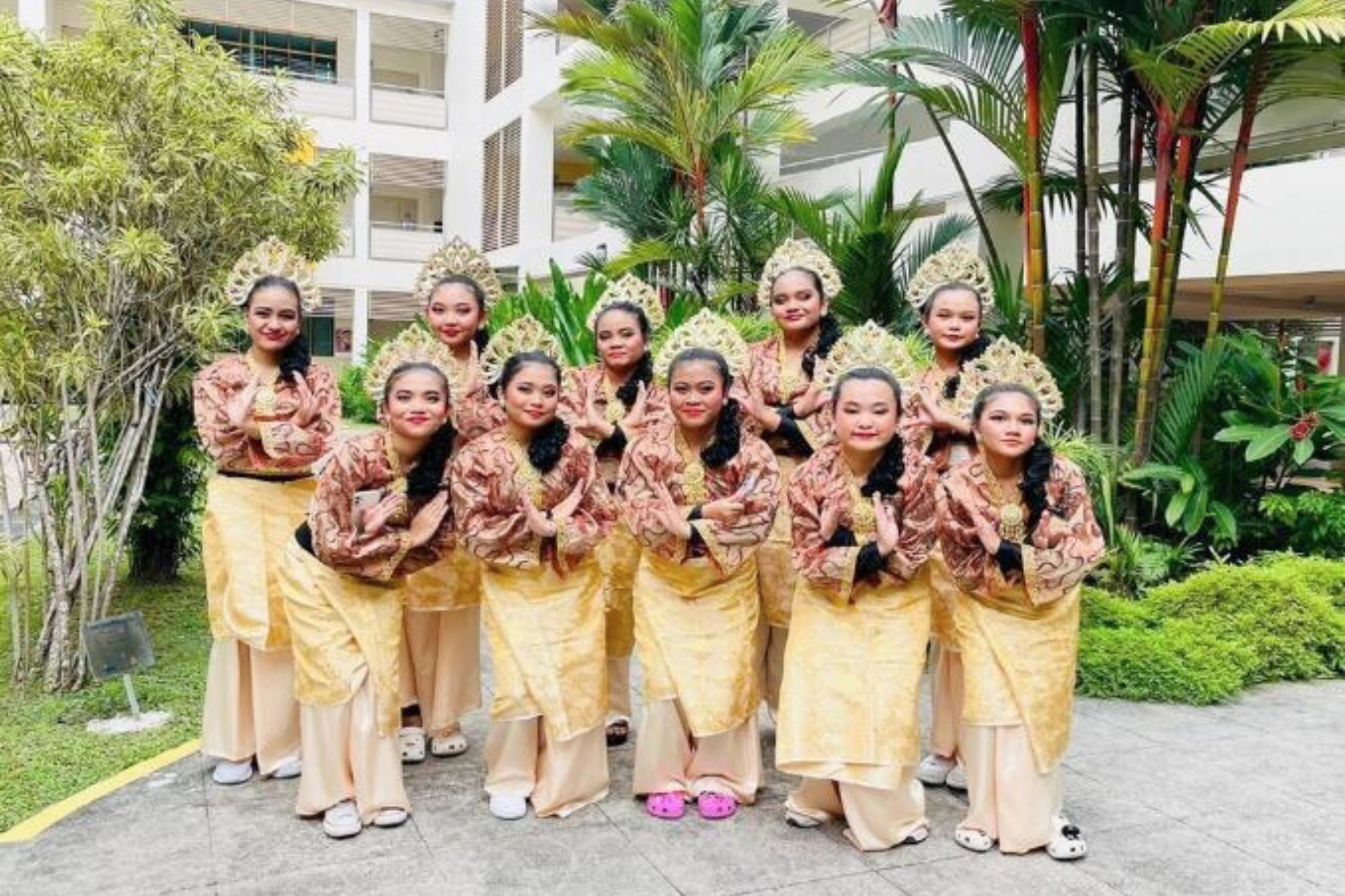 Dancers in gold costumes and ornate headpieces pose with hands together in a garden courtyard.