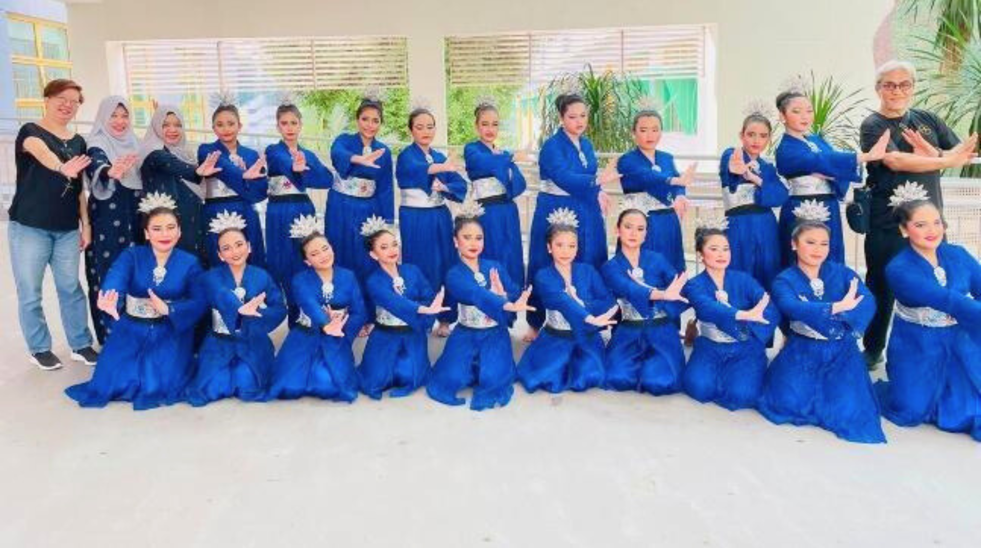 A large group of female dancers in matching royal blue traditional costumes and silver crowns posing in two rows.