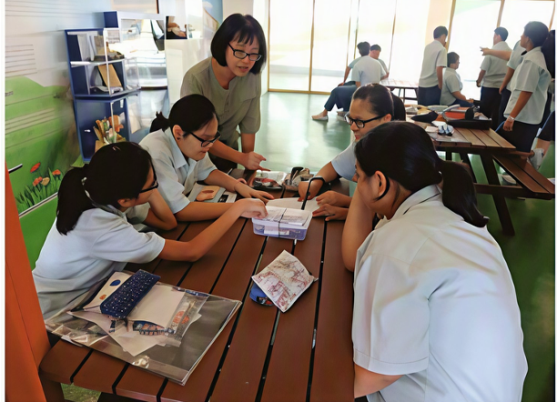 A teacher standing beside a wooden picnic table, guiding four female students in school uniform as they collaborate on a group learning activity with study materials spread out before them.