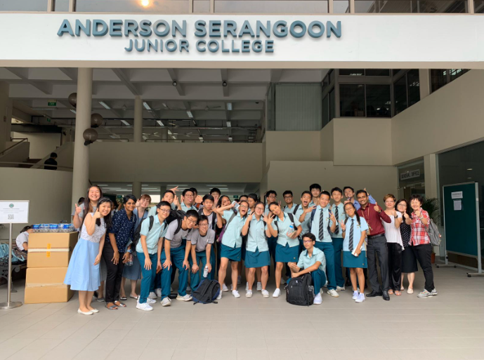 A large group of students in teal uniforms pose outside Anderson Serangoon Junior College building.