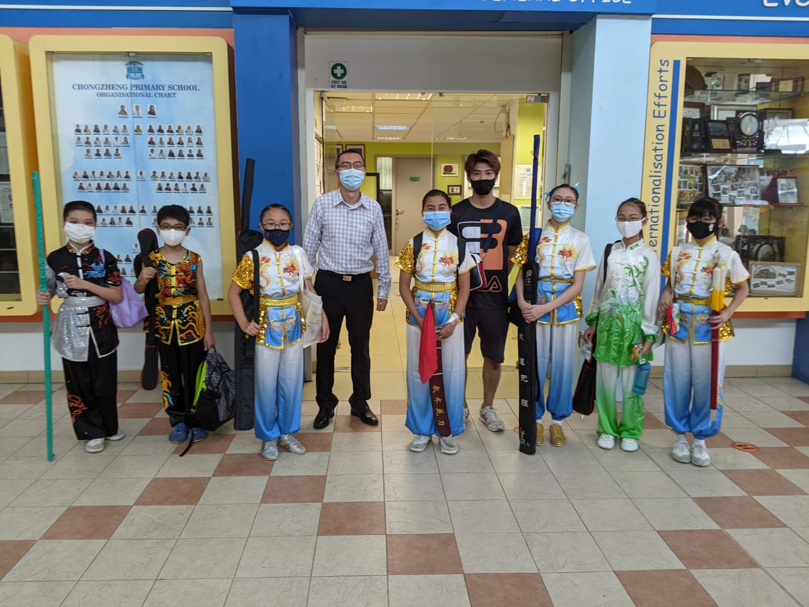 Eight masked people in wushu uniforms and two adults pose outside Chongzheng Primary School holding martial arts weapons.