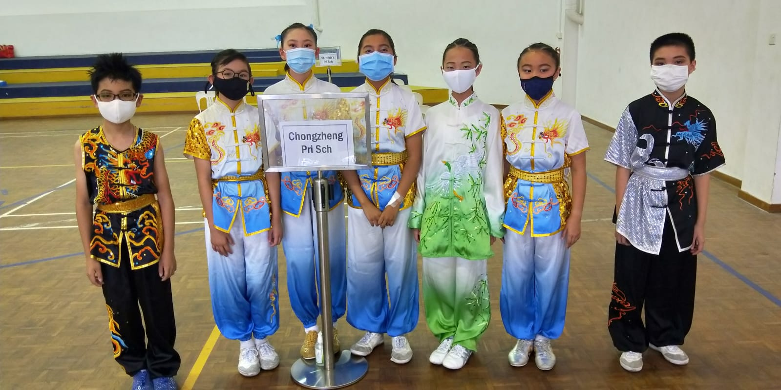 Seven children in colorful wushu costumes and face masks stand together near a Chongzheng Pri Sch sign.