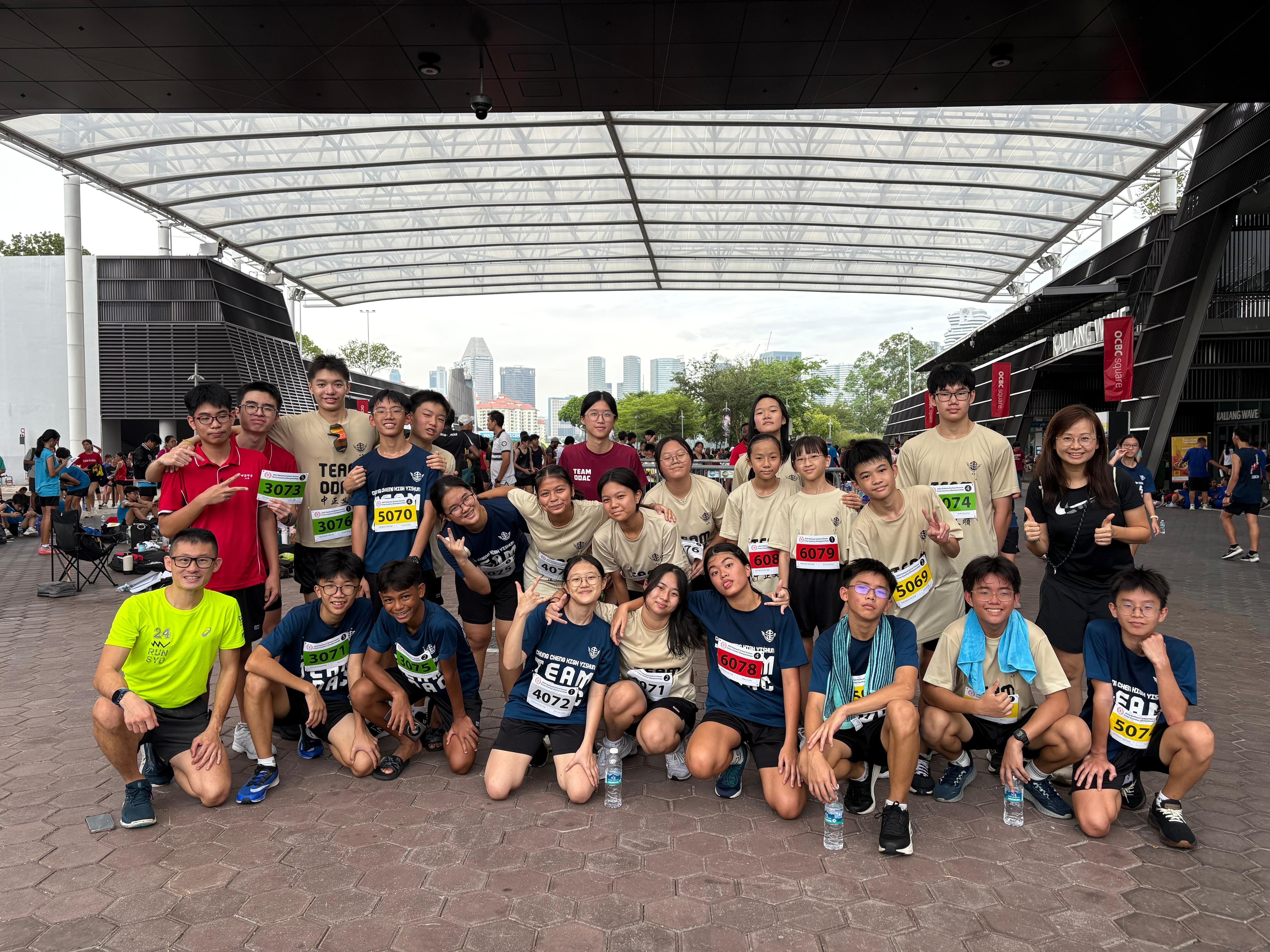 A large group of young runners wearing race bibs pose together outdoors at a running event near a stadium.